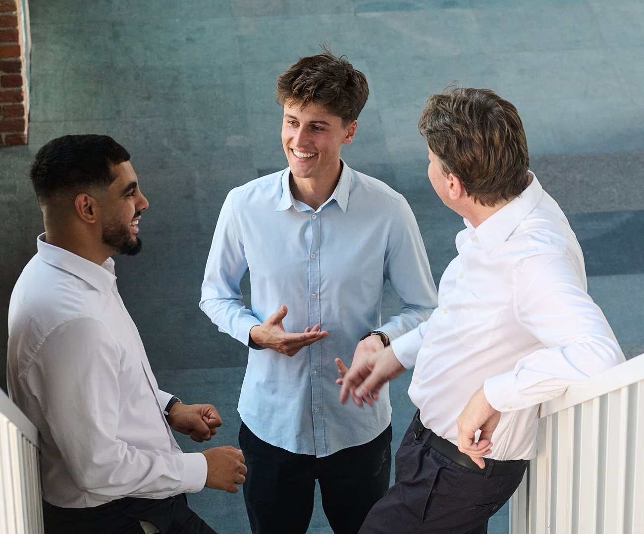 Trois hommes en chemise ayant une conversation souriante et animée dans un couloir de bureau.