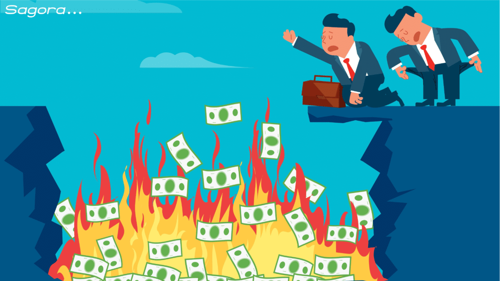 Two businessmen in suits are standing on the edge of a cliff. They look distressed as they see one-dollar notes engulfed in flames at the bottom of the ravine. One man is holding a briefcase and the other is bent over. The sky is clear with a few clouds in the background.